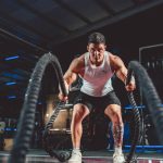Focused man in white tank top performing battle rope exercise in a modern gym.